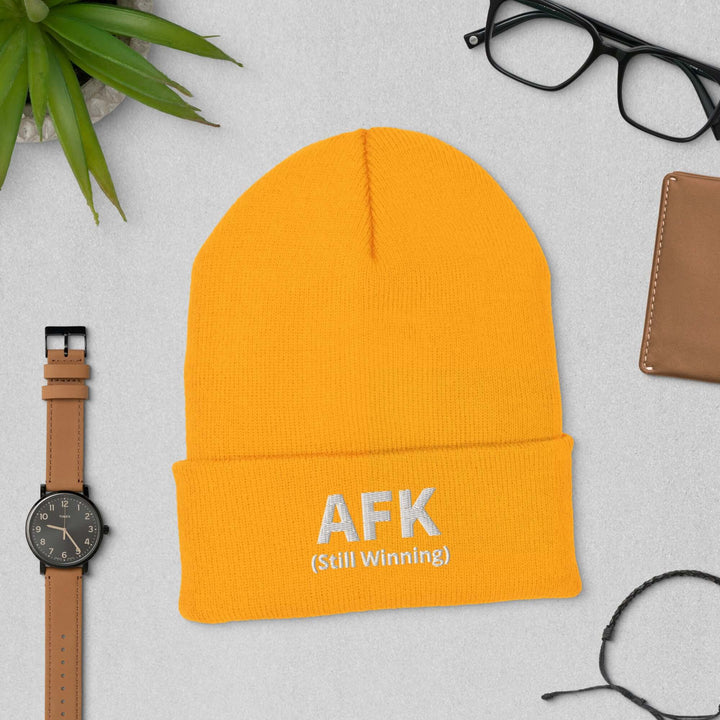 AFK Cuffed Beanie in vibrant orange with embroidered text AFK (Still Winning) on a table with accessories.