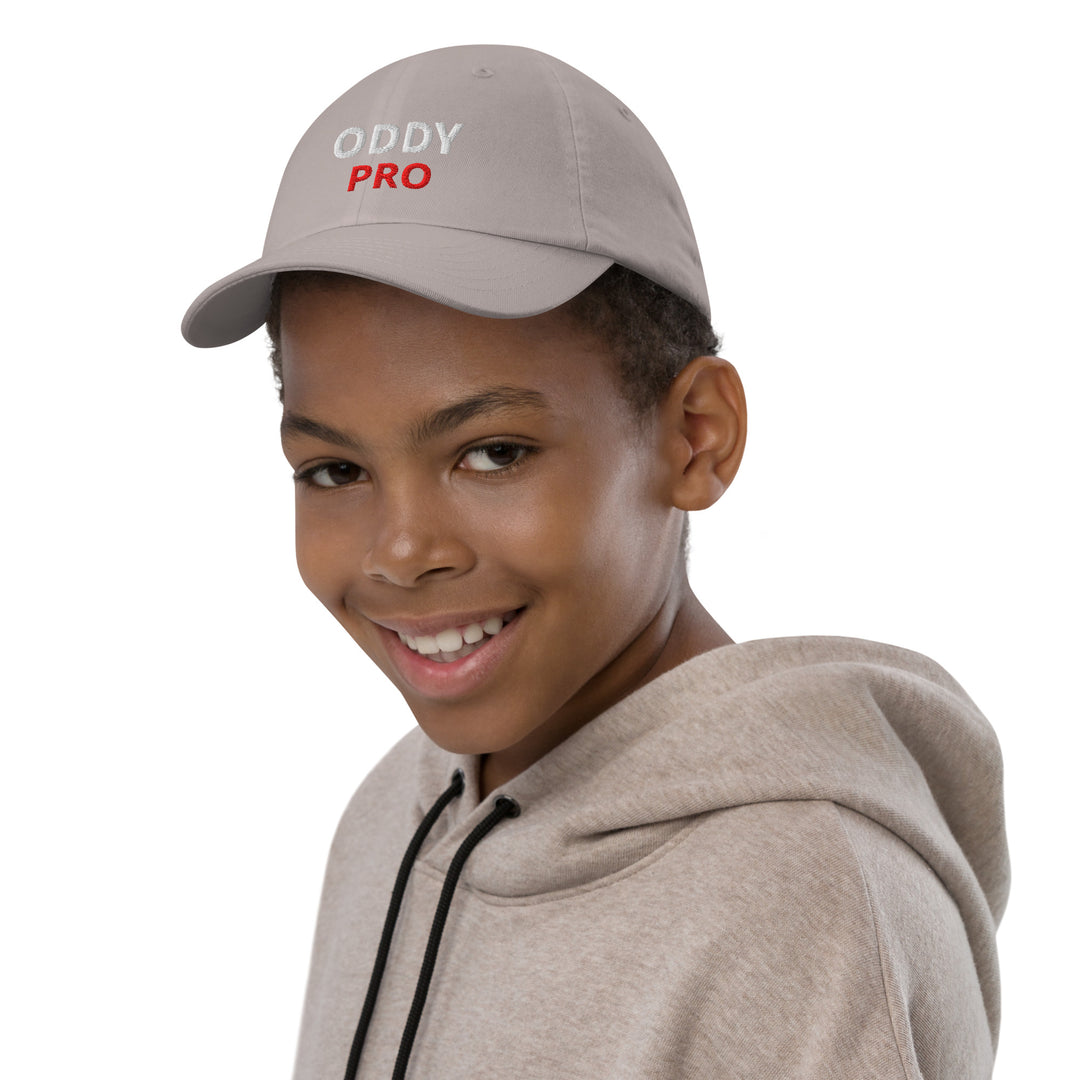 A young boy wearing the Oddy Pro Youth baseball cap, smiling, in a grey hoodie.